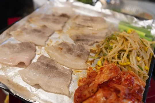 은박 불판에서 익어 가는 냉동삼겹살, 한쪽에 대파·콩나물무침·김치