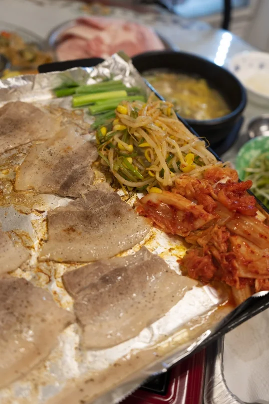 후추를 뿌려 굽는 냉동삼겹살과 대파·콩나물무침·김치, 뒤로 돌솥 국과 생고기 접시