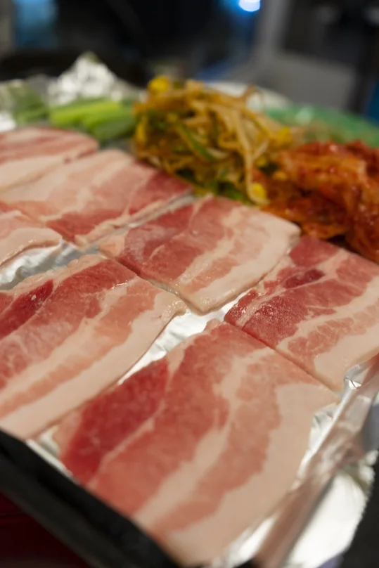 은박을 깐 불판 위에 펼친 생 냉동삼겹살, 뒤로 콩나물무침·김치·대파
