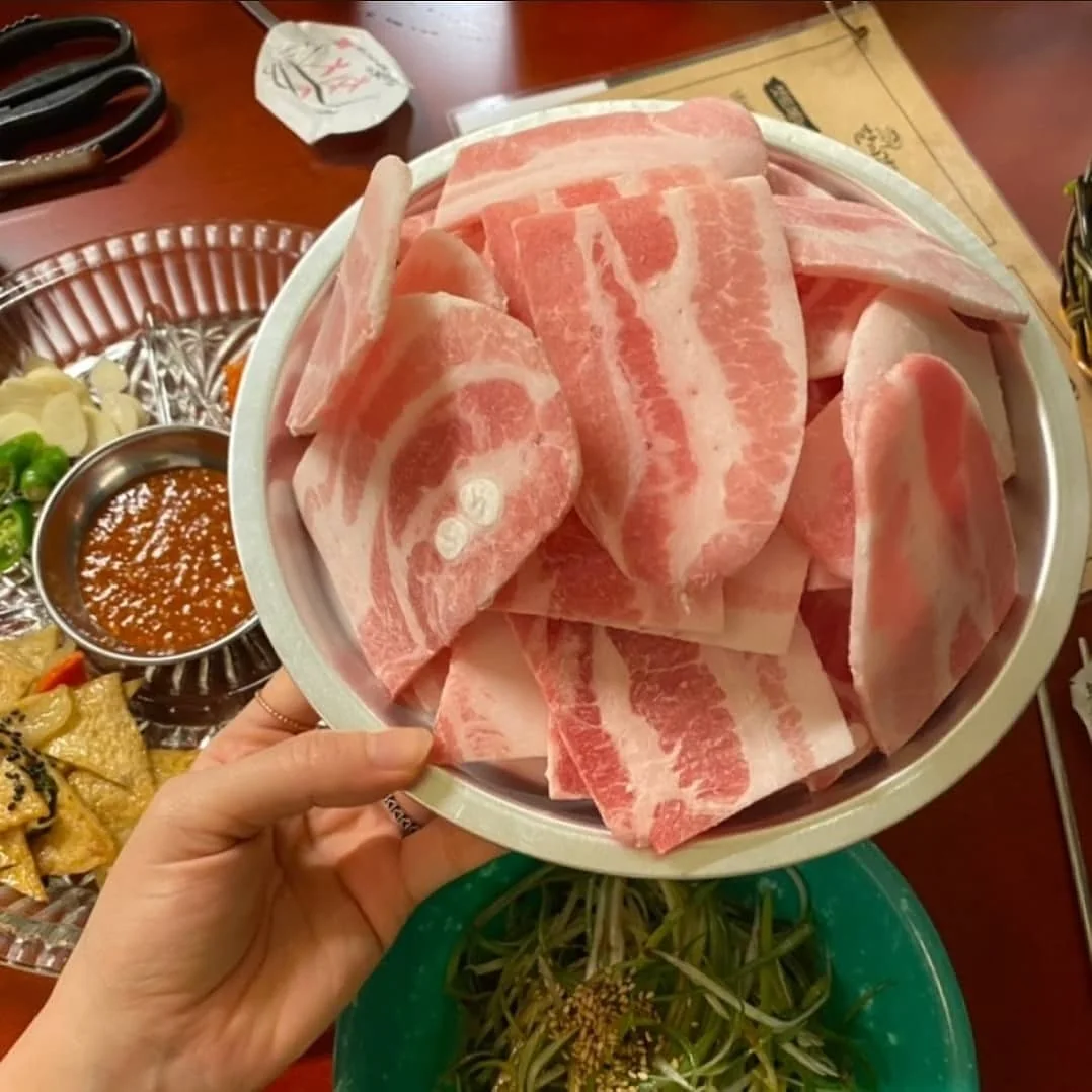 손에 든 스테인리스 접시 가득 담긴 생 냉동삼겹살, 아래로 파채 그릇과 마늘·고추·양념장·어묵 반찬