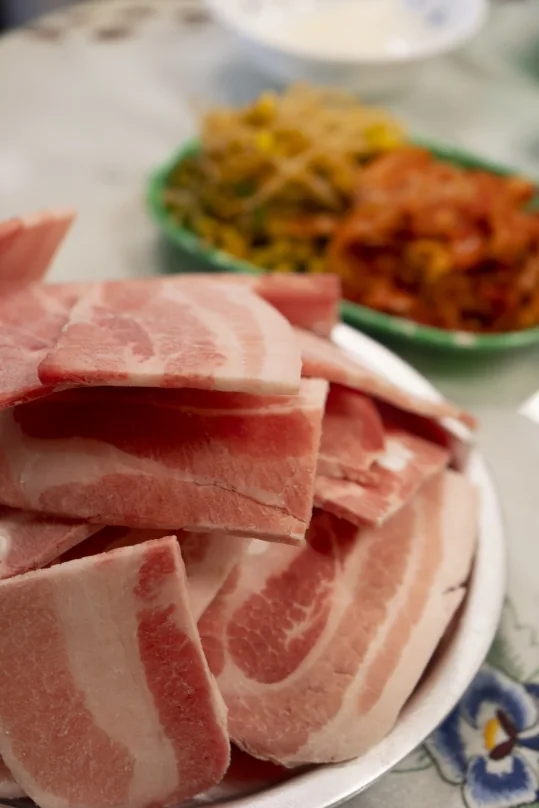 접시에 수북이 담은 생 냉동삼겹살, 뒤로 흐리게 보이는 콩나물무침과 김치, 꽃무늬 식탁보