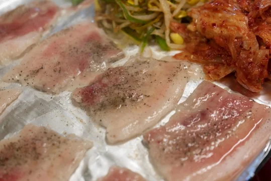 은박 불판 위 후추를 뿌린 생 냉동삼겹살 가까이, 뒤로 콩나물무침과 김치
