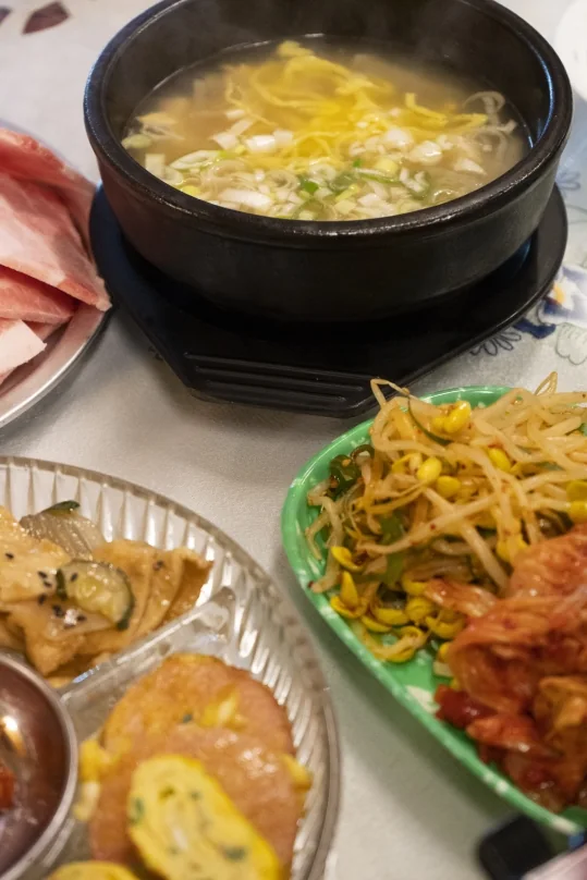 돌솥에 담긴 맑은 국과 생고기 접시, 초록 접시의 콩나물무침·김치, 유리 접시의 어묵볶음·소시지전·계란말이